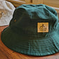 Forest green bucket hat with a tan logo on the front that reads "Falling Creek Camp" sits on a wooden bench in front of an identical hat in a tan color