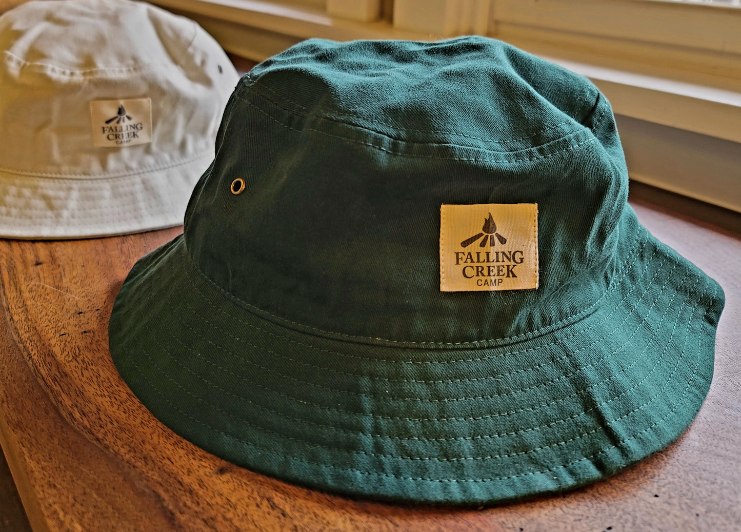Forest green bucket hat with a tan logo on the front that reads "Falling Creek Camp" sits on a wooden bench in front of an identical hat in a tan color