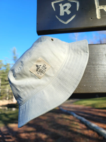 A tan putty colored bucket hat with a tan logo on the front that reads "Falling Creek Camp" hangs on the end of an outdoor wooden sign.