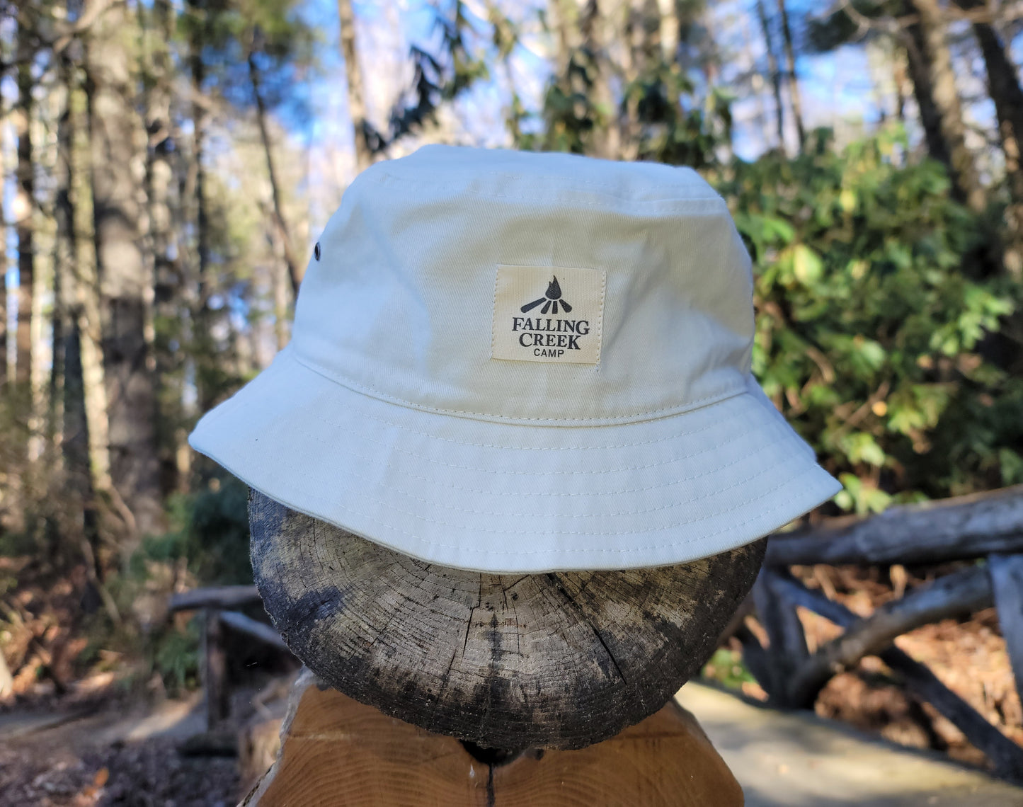 A tan putty colored bucket hat with a tan logo on the front that reads "Falling Creek Camp" sits on the end post of a wooden bridge outdoors.