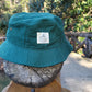 A forest green colored bucket hat with a tan logo on the front that reads "Falling Creek Camp" sits on the end post of a wooden bridge outdoors.