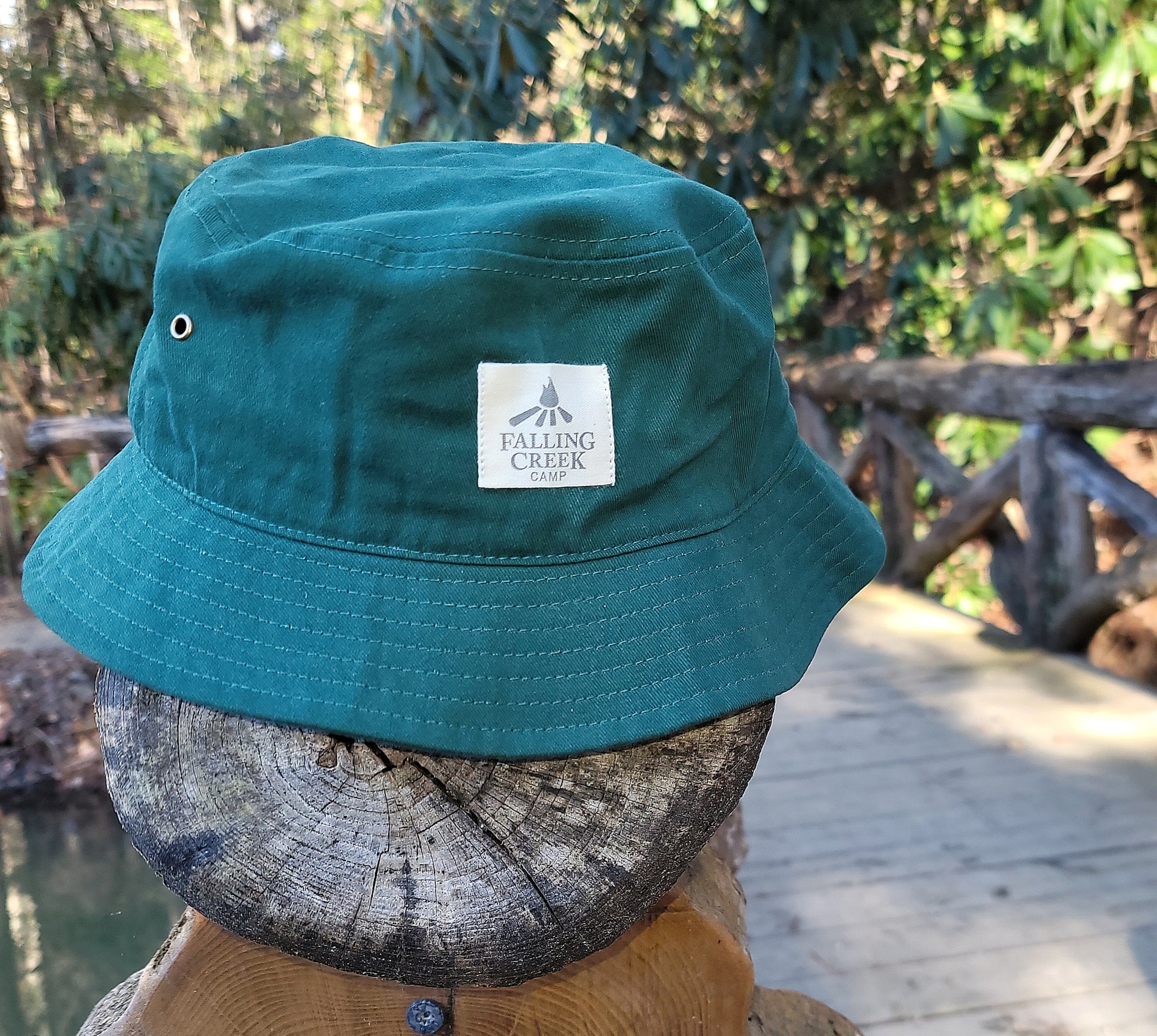 A forest green colored bucket hat with a tan logo on the front that reads "Falling Creek Camp" sits on the end post of a wooden bridge outdoors.